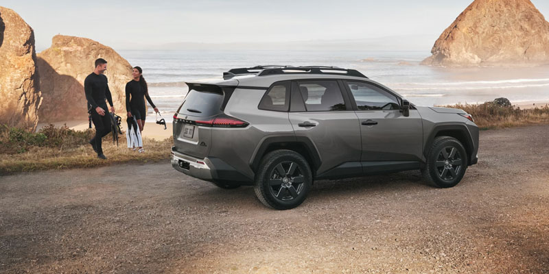 Gray Toyota RAV4 Hybrid SUV parked by a coastal beach with two people walking nearby outdoors.