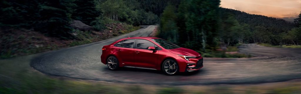 Red 2026 Toyota Corolla driving along a winding forest road at dusk, shown in motion with blurred surroundings.