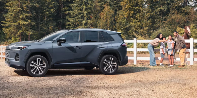 2026 Toyota RAV4 parked outdoors near a fence while a family interacts with a horse in a scenic, wooded setting.
